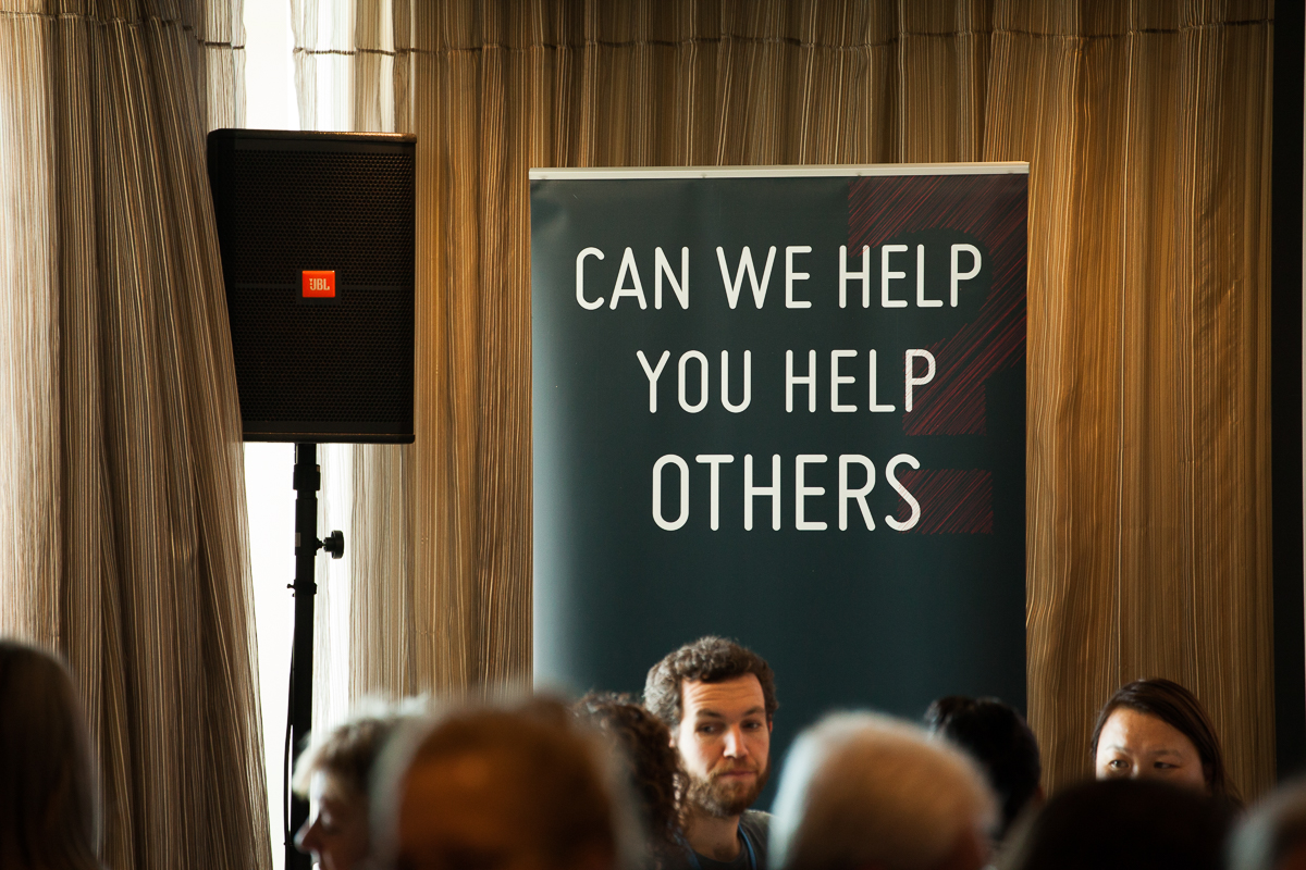 A man sitting in front of a sign that says " can we help you help others ".