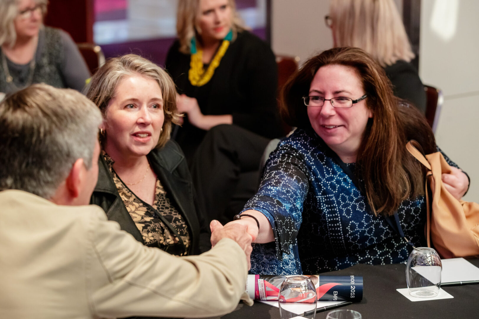 Two women shaking hands at a table with other people.